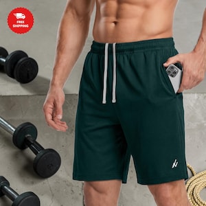 Könnte beinhalten: Dunkelgrüne Sporthose mit weißem Kordelzug und kleinem weißem Logo am Bein. Die Shorts haben Taschen. Hanteln und ein Seil sind im Hintergrund.