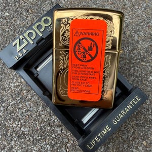 May include: A gold-colored Zippo lighter with an ornate floral design, resting in a black display case. An orange warning label is affixed to the lighter, with text that reads "Keep away from children". The display case has the text "Lifetime Guarantee".
