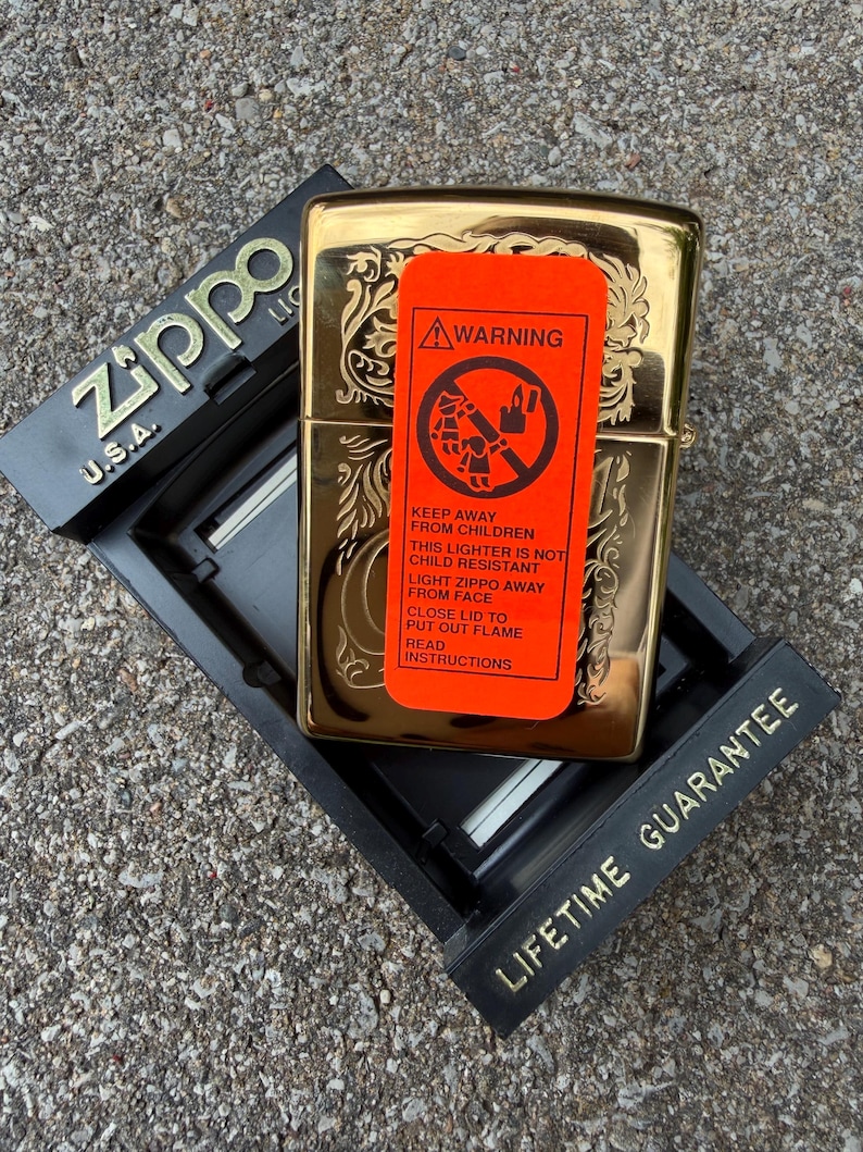 May include: A gold-colored Zippo lighter with an ornate floral design, resting in a black display case. An orange warning label is affixed to the lighter, with text that reads "Keep away from children". The display case has the text "Lifetime Guarantee".