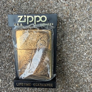 May include: A gold-colored Zippo lighter with an ornate floral design, packaged in a black box. The box features the Zippo logo and the text "U.S.A. LIGHTER". The packaging also states "LIFETIME GUARANTEE".