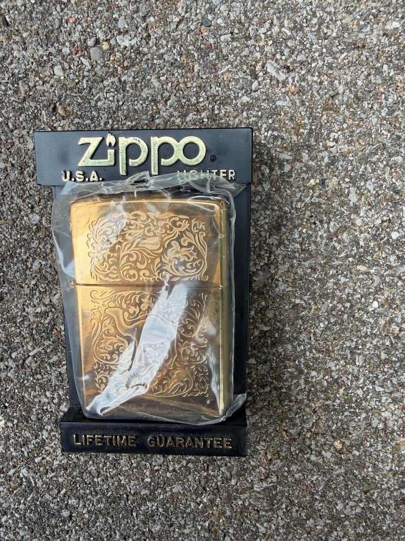 May include: A gold-colored Zippo lighter with an ornate floral design, packaged in a black box. The box features the Zippo logo and the text "U.S.A. LIGHTER". The packaging also states "LIFETIME GUARANTEE".