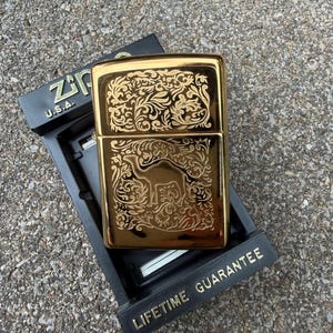 May include: A gold-colored Zippo lighter with an ornate floral and camel design. The lighter is in its original black box, which has the text "Zippo U.S.A." and "Lifetime Guarantee" in gold lettering.