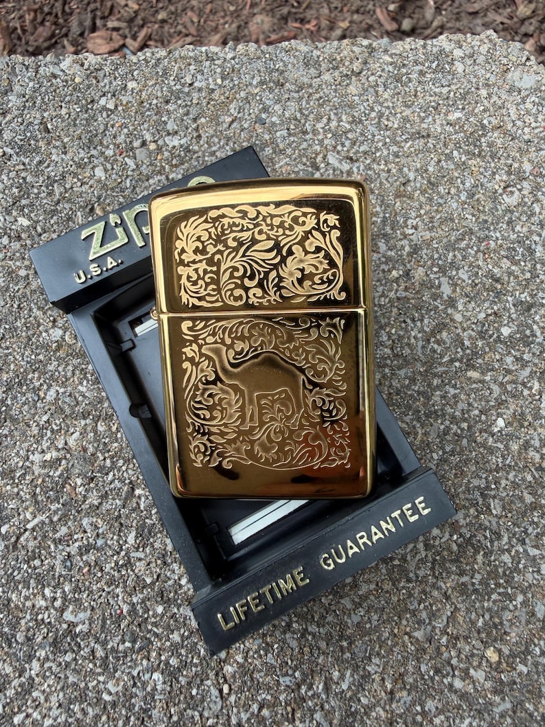 May include: A gold-colored Zippo lighter with an ornate floral and camel design. The lighter is in its original black box, which has the text "Zippo U.S.A." and "Lifetime Guarantee" in gold lettering.