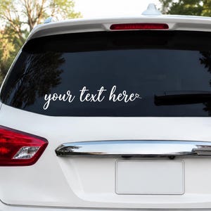 May include: A white vinyl decal applied to the rear window of a white vehicle. The decal features the text your text here in a cursive script font, with a small heart icon integrated into the end of the final word of the phrase.