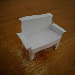 May include: A small, white paper bench with a back and armrests. The miniature furniture piece is constructed from folded paper and sits on a wooden surface. The bench is a simple, handcrafted item.