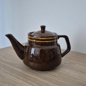 May include: A glossy dark brown ceramic teapot with a matching lid. The body features a wide, rounded base that tapers toward the top, accented by two horizontal light brown stripes encircling the upper section near the lid and a curved handle.