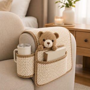 May include: A cream-colored crochet sofa arm organizer with a brown trim. It features a cup holder, a pocket with a book, and a teddy bear. The organizer is designed to fit over a sofa arm.