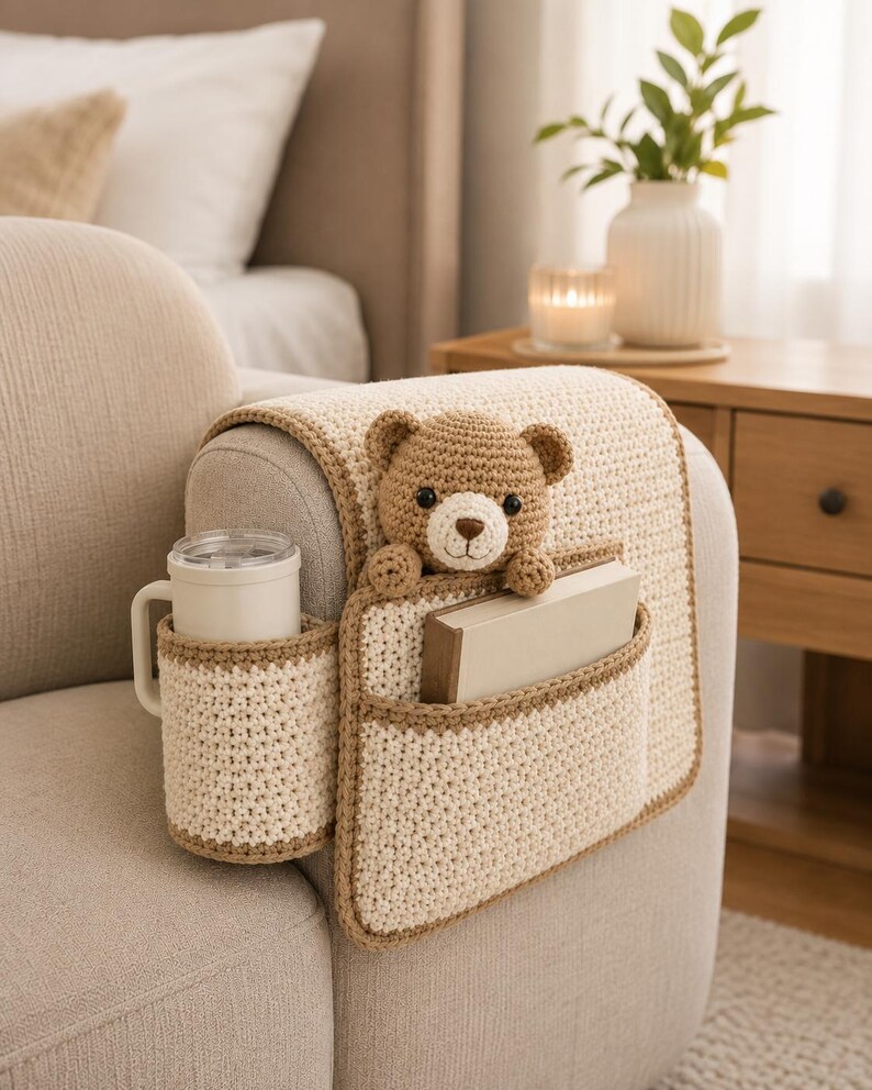 May include: A cream-colored crochet sofa arm organizer with a brown trim. It features a cup holder, a pocket with a book, and a teddy bear. The organizer is designed to fit over a sofa arm.