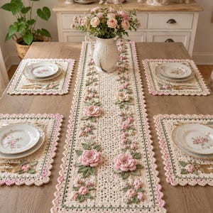 Puede incluir: Un juego de mesa de comedor con un camino de mesa y manteles individuales de ganchillo color crema. El camino de mesa y los manteles individuales están adornados con rosas rosas y hojas verdes. Cada cubierto incluye un plato blanco con un diseño floral, un tenedor y un cuchillo.