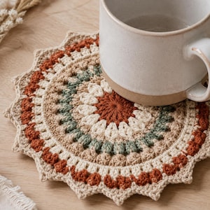 May include: A handmade crochet coaster with a circular design. The coaster features a mix of colors, including beige, rust, green, and cream. A white ceramic mug sits on top of the coaster, adding a touch of contrast to the warm color palette.