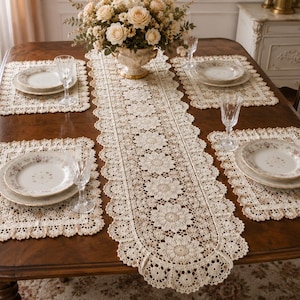 May include: A formal dining table set for four, featuring a dark wood table with a cream-colored lace runner and matching placemats. Each place setting includes a floral-patterned plate, a water glass, and a bouquet of flowers in a vase.