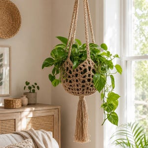 May include: A beige macrame plant hanger with a tassel, holding a lush green plant with variegated leaves. The plant hanger is suspended in front of a window, showcasing a natural, bohemian aesthetic. The woven design adds texture and visual interest.