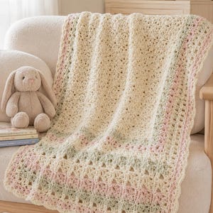 May include: Cream-colored crochet baby blanket with pink and green stripes. The blanket is draped over a cream-colored armchair. A stuffed bunny and a stack of books are on the chair. The blanket has a scalloped edge.