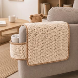 May include: A beige crochet sofa arm cover with a built-in cup holder and a small bear hat. The cover has a brown border and is placed on a gray sofa. The bear hat is also beige.