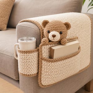 May include: A crocheted sofa armrest organizer in cream and brown. It features a cup holder, a pocket holding a book, and a teddy bear. The organizer is designed to fit over the arm of a sofa.