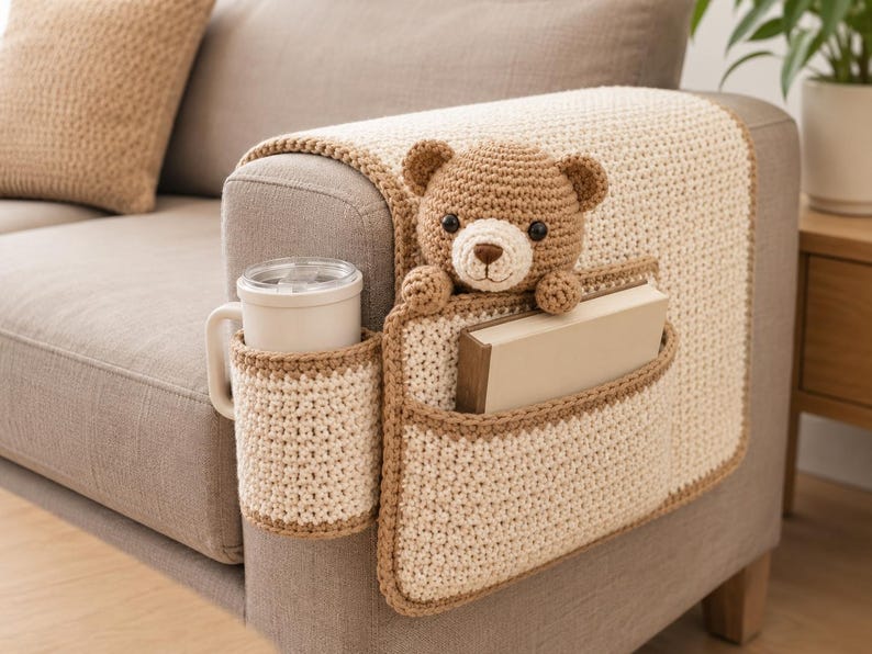 May include: A crocheted sofa armrest organizer in cream and brown. It features a cup holder, a pocket holding a book, and a teddy bear. The organizer is designed to fit over the arm of a sofa.