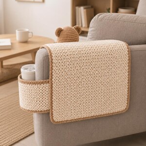 May include: A beige crochet sofa arm cover with a built-in cup holder and a brown trim. A small, brown crochet bear hat sits on top of the arm cover. The cover is placed on a gray sofa.