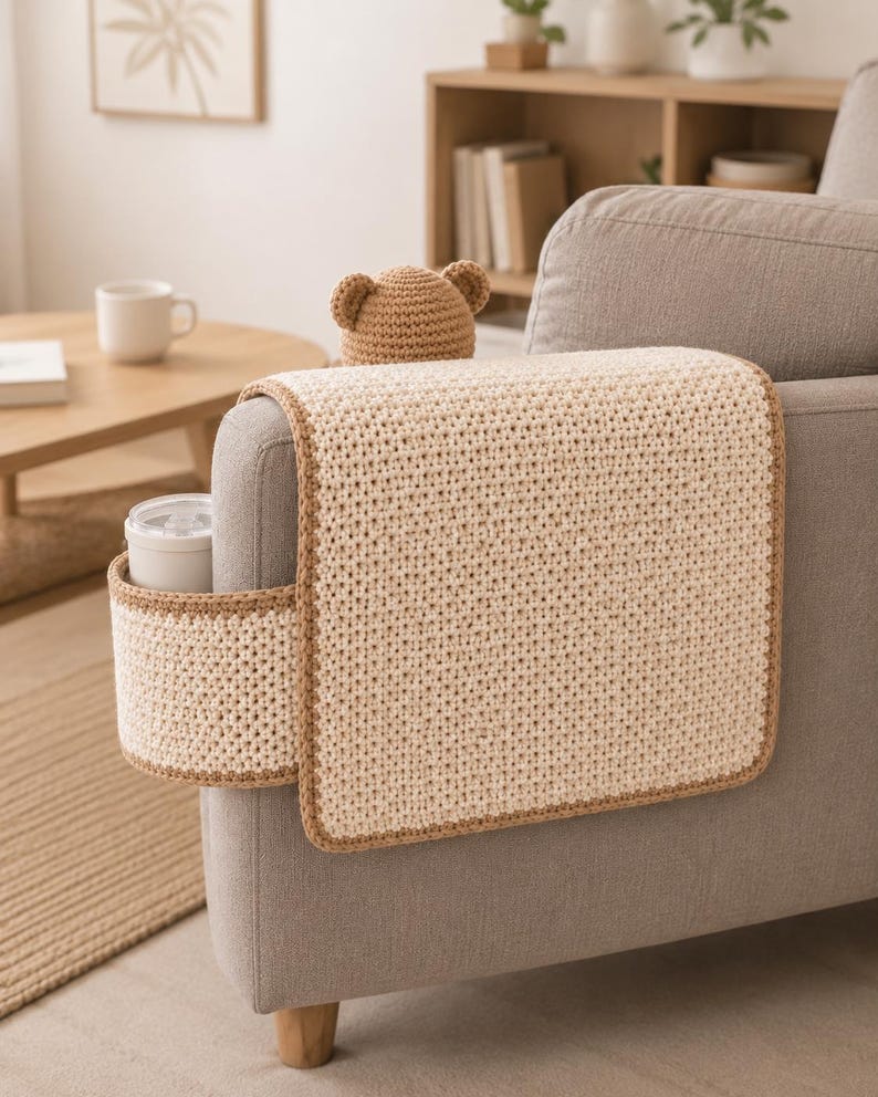 May include: A beige crochet sofa arm cover with a built-in cup holder and a brown trim. A small, brown crochet bear hat sits on top of the arm cover. The cover is placed on a gray sofa.
