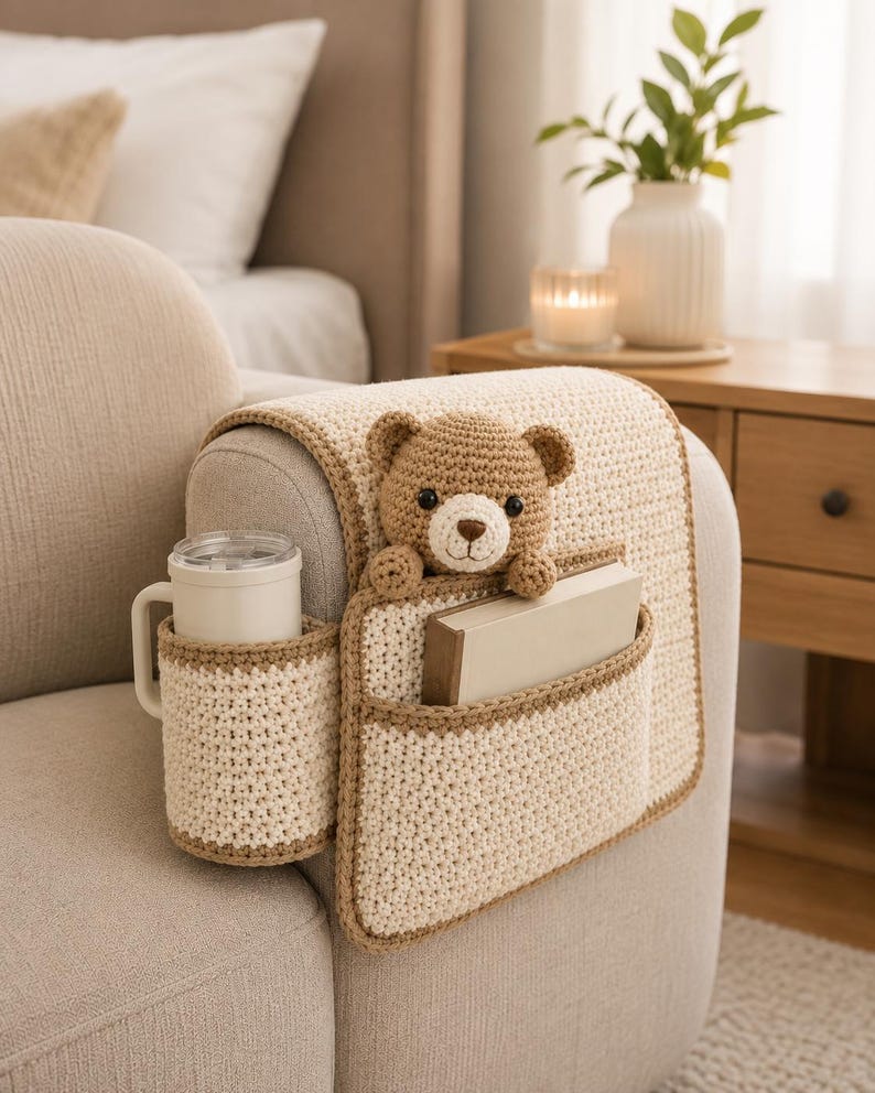 May include: A cream-colored crochet sofa arm organizer with a brown trim. It features a cup holder, a pocket with a book, and a teddy bear. The organizer is designed to fit over the arm of a sofa.