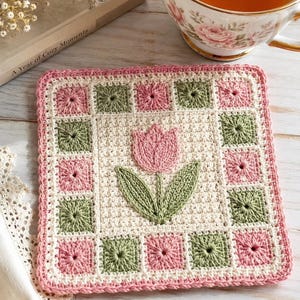 May include: A square crochet coaster featuring a central cream-coloured panel with a pink tulip and green leaves. The border is composed of alternating pink and sage green square motifs, finished with a pink crocheted edge around the perimeter.