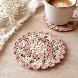May include: Handmade crochet coasters in a floral design. The coasters feature a scalloped edge and a color palette of pink, cream, and green. One coaster has a coffee cup on top. The coasters are approximately 4 inches in diameter.