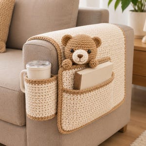 May include: A crocheted sofa armrest organizer in cream and brown. It features a cup holder, a pocket for a book, and a crocheted teddy bear. The organizer is placed on a beige sofa, with a matching pillow visible.