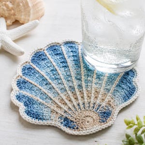 May include: A round crochet coaster shaped like a seashell, featuring a gradient of blue and beige yarn. The design includes radiating lines that mimic the texture of a scallop shell, finished with a solid cream-colored border around the edge.