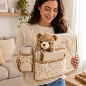 May include: A crocheted organizer in cream and brown, featuring pockets for a mug, books, and a teddy bear. The handmade item is designed to hold various items. The teddy bear is brown and has a cream-colored snout.