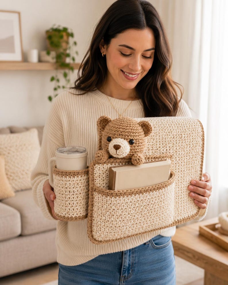 May include: A crocheted organizer in cream and brown, featuring pockets for a mug, books, and a teddy bear. The handmade item is designed to hold various items. The teddy bear is brown and has a cream-colored snout.