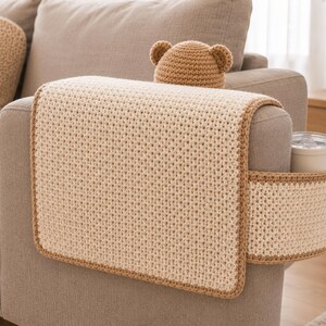 May include: A beige crochet sofa arm cover with a matching cup holder and a bear-eared hat. The cover has a brown border and is placed on a gray sofa arm. A white mug is in the cup holder.