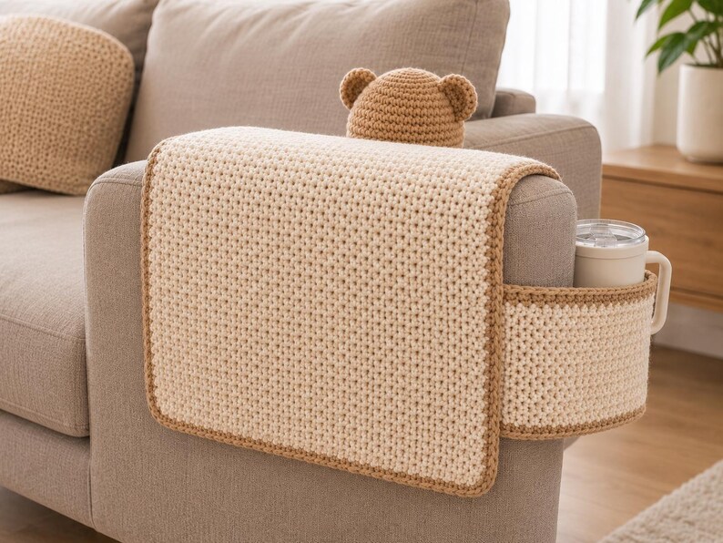 May include: A beige crochet sofa arm cover with a matching cup holder and a bear-eared hat. The cover has a brown border and is placed on a gray sofa arm. A white mug is in the cup holder.