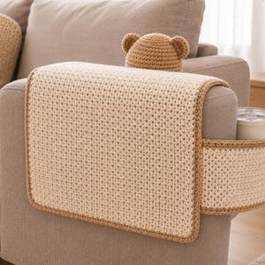 May include: A beige crochet sofa arm cover with a matching cup holder and a bear-eared hat. The cover has a brown border and is placed on a gray sofa arm. A white mug is in the cup holder.
