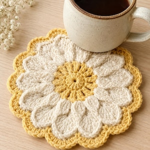 May include: A handmade crochet coaster in a daisy flower design. The coaster features a yellow center and scalloped edge, with white petals. A ceramic mug filled with coffee sits on the coaster.