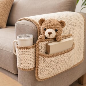 May include: A crocheted sofa armrest organizer in cream and brown. It features a cup holder, a pocket holding a book, and a teddy bear. The organizer is designed to fit over the arm of a sofa.