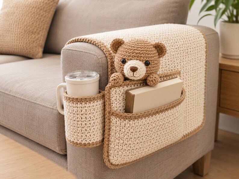 May include: A crocheted sofa armrest organizer in cream and brown. It features a cup holder, a pocket holding a book, and a teddy bear. The organizer is designed to fit over the arm of a sofa.