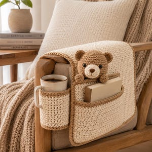 May include: Crocheted armrest organizer in cream and brown, featuring a cup holder, a pocket with a book, and a teddy bear. The organizer is placed on a wooden chair arm, with a beige pillow and blanket in the background.