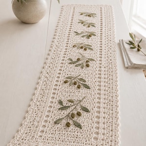 May include: A rectangular cream-colored crochet table runner featuring a central row of embroidered olive branches with green leaves and small brown olives. The runner has a decorative scalloped edge and an intricate openwork border pattern.