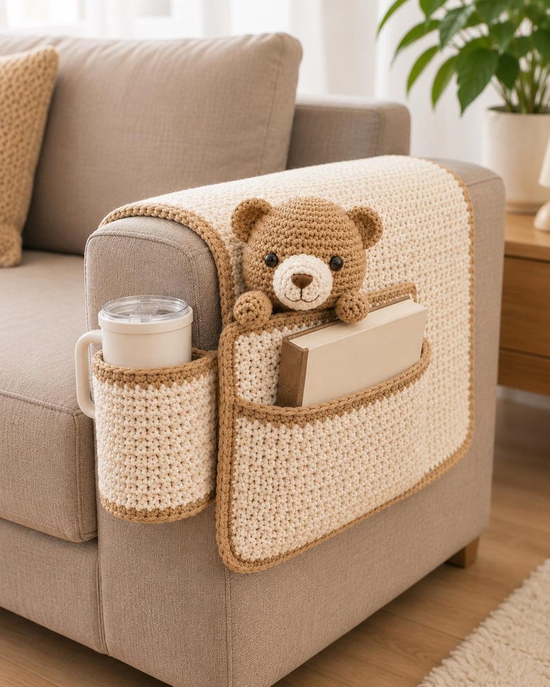 May include: A crocheted sofa armrest organizer in cream and brown. It features a cup holder, a pocket for a book, and a crocheted teddy bear. The organizer is placed on a beige sofa, with a matching pillow visible.