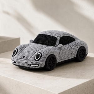 May include: A handcrafted crochet toy car in a light grey colour with black accents. The vehicle features a rounded body, black circular wheels, black windows, and circular headlights with white outlines, mimicking the shape of a sports car.