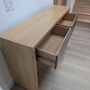 Pode incluir: Uma mesa de consola de madeira clara com duas gavetas, uma parcialmente aberta. A mesa é feita de madeira clara e tem um design simples e moderno. As gavetas são de um tom castanho mais escuro. A mesa está colocada contra uma parede branca.