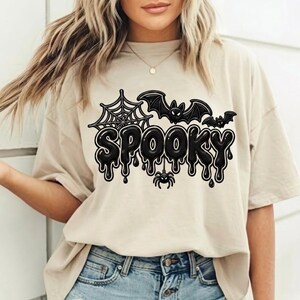 May include: A beige short-sleeve t-shirt featuring a black graphic print with the word SPOOKY in a dripping font. The design includes a spider web, two bats, and a small spider hanging from the letters, all rendered in a bold black outline.