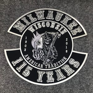 May include: A black and white embroidered patch featuring a grim reaper holding a playing card with an ace of spades. The text reads MILWAUKEE WISCONSIN, 1903, 2018, AMERICAN TRADITION, and 115 YEARS. The design is circular with arched top and bottom sections.