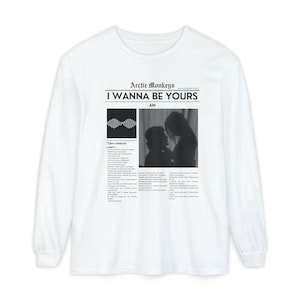以下が含まれることがあります： 白の長袖Tシャツ。黒字で「Arctic Monkeys I Wanna Be Yours」と書かれています。デザインには白黒画像と歌詞が含まれています。Tシャツはクルーネックです。