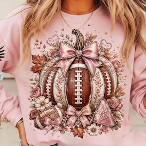 May include: A light pink sweatshirt featuring a central graphic of a pumpkin shaped like a football, adorned with pink satin bows, glittery accents, autumn leaves, and roses. A smaller football and a pennant with the text GAME DAY are included.