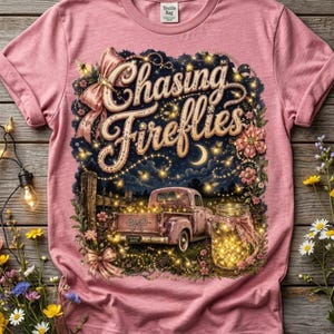 May include: A dusty pink t-shirt featuring a graphic print of a vintage pickup truck, a mason jar filled with glowing lights, and the text Chasing Fireflies. The design includes floral accents, bows, and a crescent moon against a dark night sky.