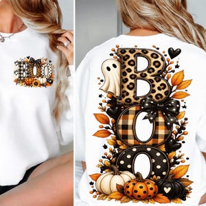 May include: A white sweatshirt featuring the text BOO in large letters. The B has a leopard print, the O has a buffalo plaid pattern, and the second O has black and white polka dots. A ghost, pumpkins, leaves, and a black bow decorate the design.