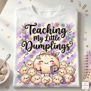 May include: A white t-shirt featuring a graphic of a large dumpling character wearing a headband and holding a book labelled A+, surrounded by smaller dumpling characters. The text reads Teaching My Little Dumplings in black script.