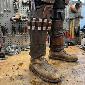 K&ouml;nnte beinhalten: Ein Paar abgenutzte braune Lederstiefel mit dunkelbraunen Lederschienbeinschonern. Die Schoner sind mit einem horizontalen Lederriemen verziert, der sechs metallische zylindrische Kanister h&auml;lt, sowie einer vertikalen Tasche mit einem blauen Werkzeug.