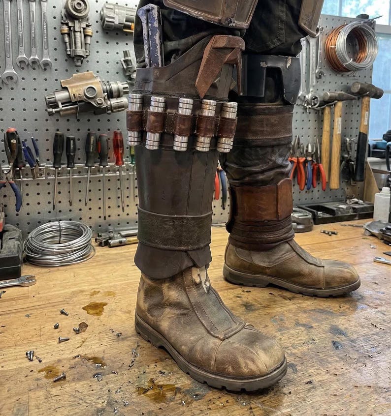 K&ouml;nnte beinhalten: Ein Paar abgenutzte braune Lederstiefel mit dunkelbraunen Lederschienbeinschonern. Die Schoner sind mit einem horizontalen Lederriemen verziert, der sechs metallische zylindrische Kanister h&auml;lt, sowie einer vertikalen Tasche mit einem blauen Werkzeug.