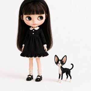 May include: A doll with long dark hair, a black headband, and a black dress with a white collar and cuffs. The doll wears beige socks and black Mary Jane shoes. Beside the doll stands a small black and white cat figurine with large ears.